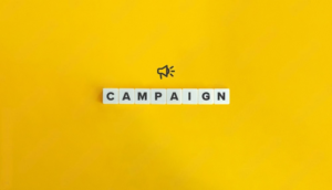 Marketing campaign concept with word 'Campaign' spelled out in letter tiles on a bright yellow background, accompanied by a megaphone icon symbolizing promotion and advertising.